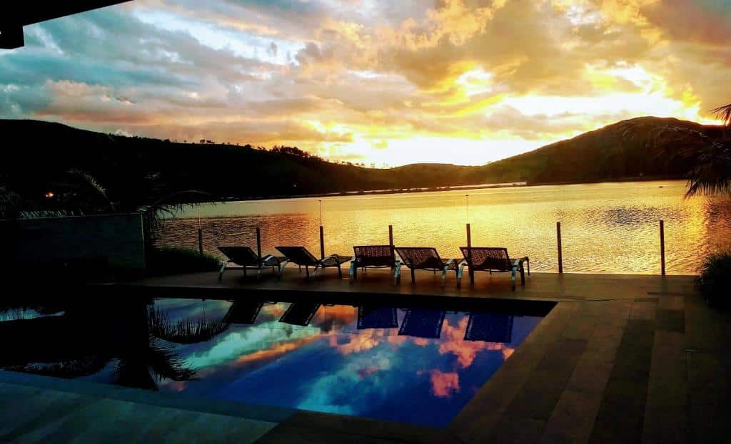 Pôr do sol em um lago calmo com espreguiçadeiras à beira de uma piscina, refletindo o céu colorido e as colinas ao redor, do Cyrilo's Palace Hotel. Representa hotéis em Capitólio.