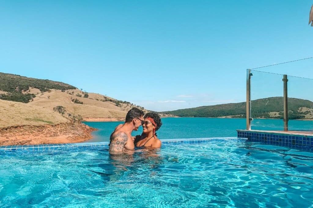 Um casal está junto em uma piscina com colinas e um lago ao fundo, sob um céu azul claro, do Hotel Boutique Coração Aberto. Representa hotéis em Capitólio.
