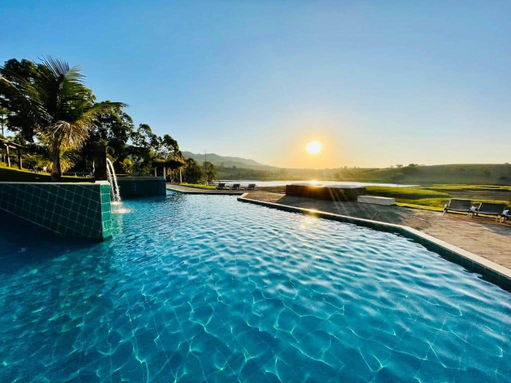 Piscina infinita com elementos aquáticos ao nascer do sol, com vista para campos gramados e colinas distantes sob um céu claro, do Hotel Chalé Capitólio.