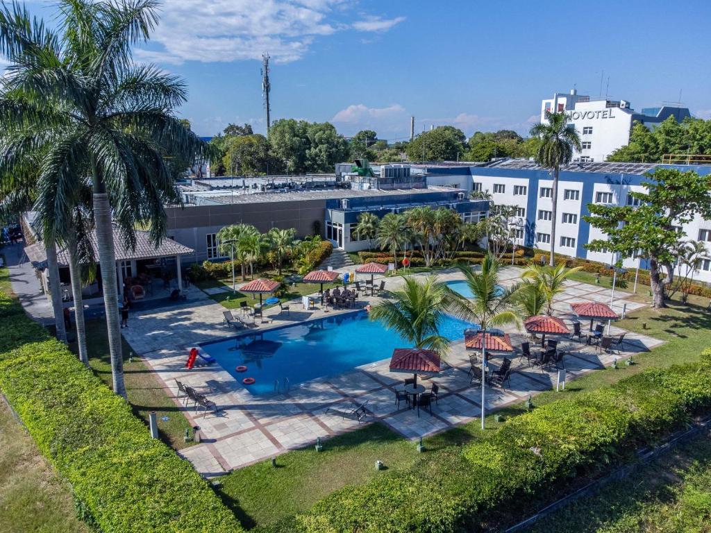 A área da piscina de um hotel com espreguiçadeiras, guarda-sóis, palmeiras e um edifício do Novotel Manaus ao fundo em um dia ensolarado. Representa hotéis em Manaus.