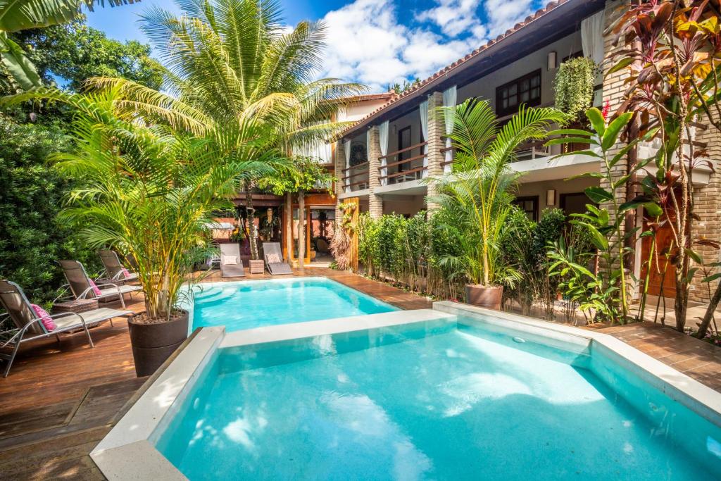 Piscina externa da Pousada Água de Coco cercada por palmeiras e espreguiçadeiras, ao lado de um prédio de dois andares com varandas e vegetação. Representa hotéis em Arraial d’Ajuda.