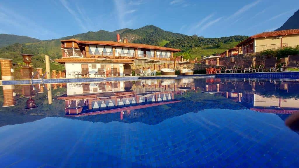 Uma casa grande e colinas verdes são refletidas na água azul de uma piscina externa em um dia ensolarado, da Pousada Aldeia Manacás. Representa hotéis em São Bento do Sapucaí.