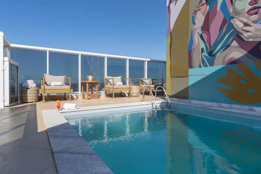 Pequena piscina externa do Royal Urban Macaé Hotel com espreguiçadeiras à beira da piscina, uma mesa e um mural colorido na parede adjacente sob um céu claro.