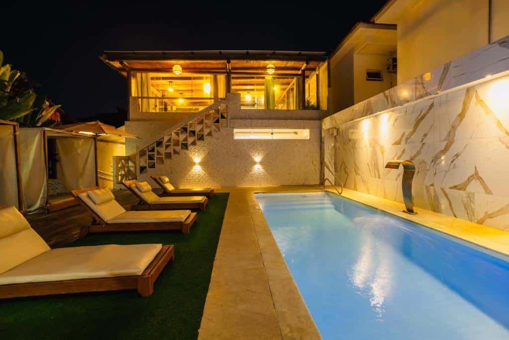 Moderna área da piscina à noite com espreguiçadeiras, paredes iluminadas e uma área de estar no andar superior com iluminação quente, do Santorini Beach Hotel. Representa hotéis em Arraial d’Ajuda.