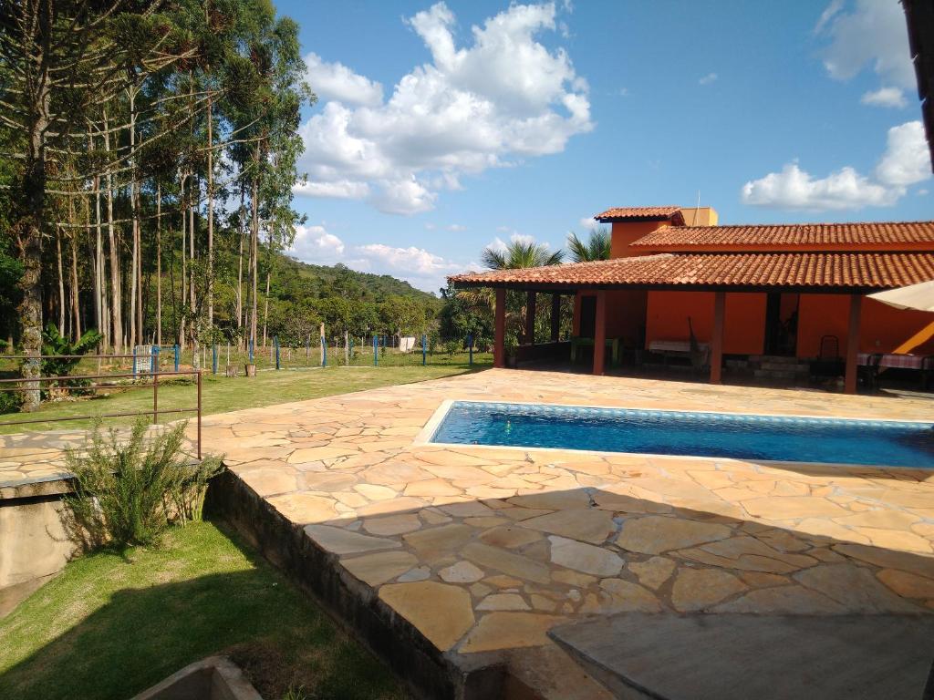 Um pátio de pedra com uma piscina ao lado de uma casa de laranja, cercado por árvores e um quintal gramado sob um céu azul, no Sítio Pé da Serra - Pousada Rural. Representa hotéis em Capitólio.