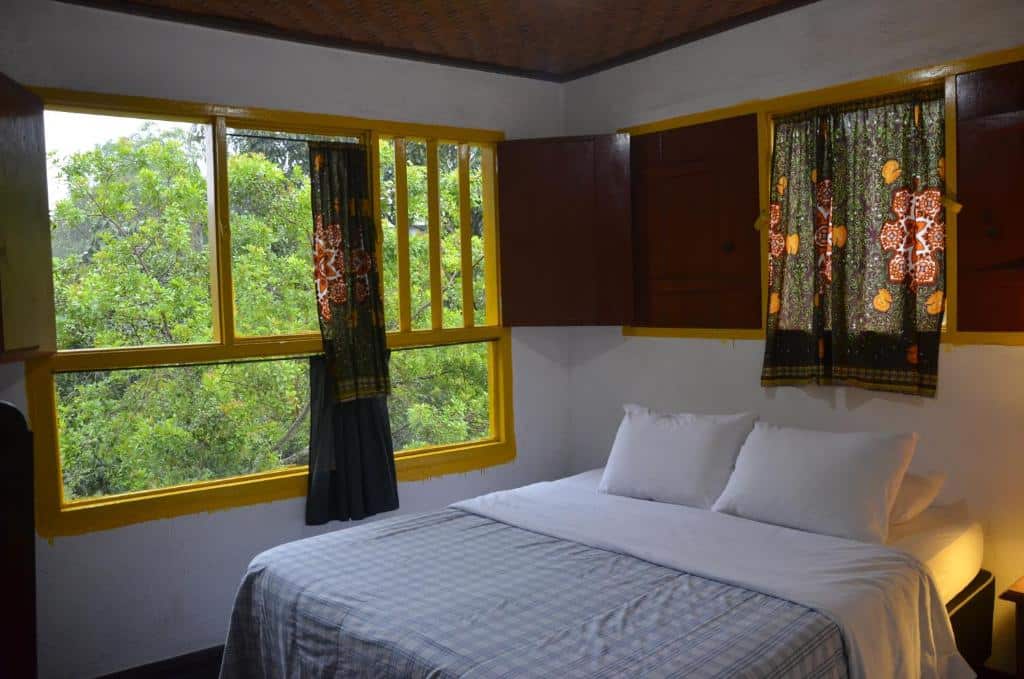 Quarto da Pouso da Passagem B&B com uma cama com lençóis brancos ao lado de grandes janelas com moldura amarela e cortinas estampadas com vista para árvores verdes.