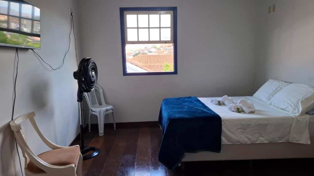 Um quarto simples do Solar do Carmo Simples com uma cama, duas cadeiras, um ventilador de pé, uma TV na parede e uma janela com vista para o exterior.