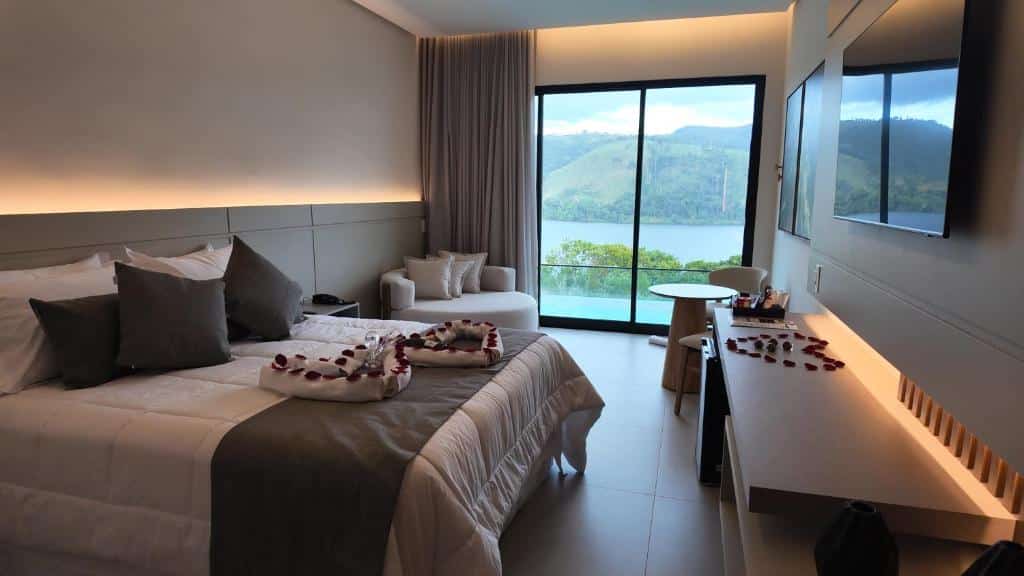 Quarto do Collegare Hotel com cama, pétalas de rosa, área de estar, escrivaninha e janela com vista para um lago e colinas.