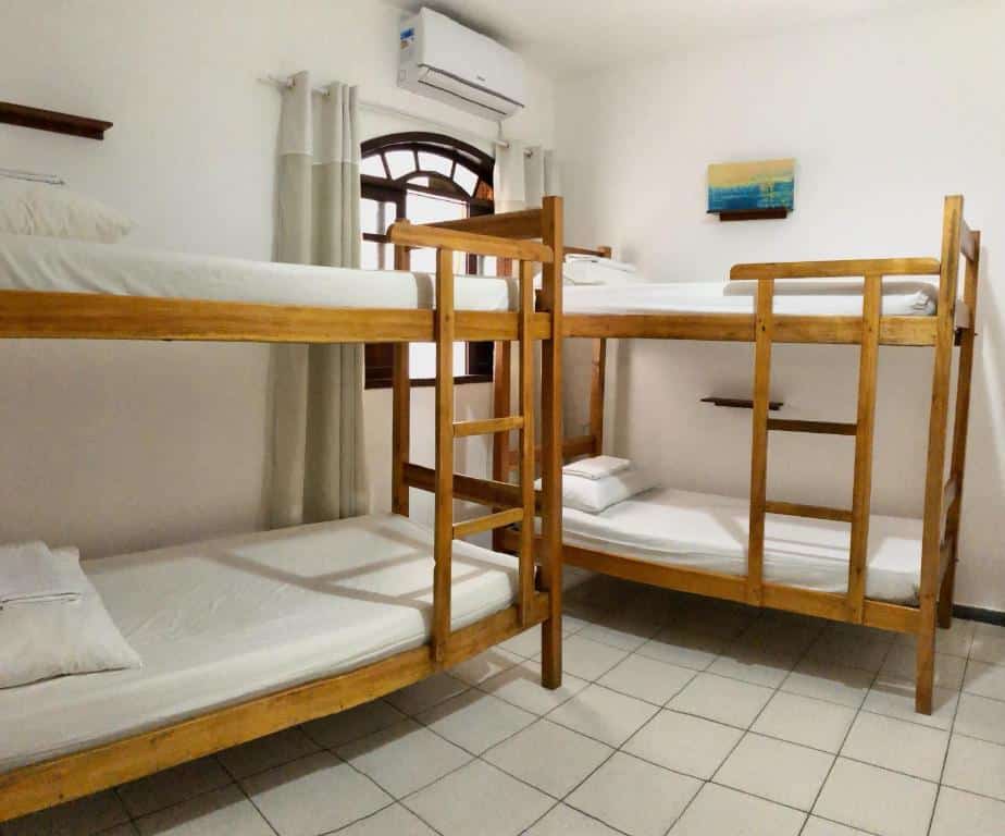 Um quarto com dois beliches de madeira, cada um com roupa de cama branca, piso de azulejo e uma janela com cortinas brancas, do Odoyá Casa Hostel.