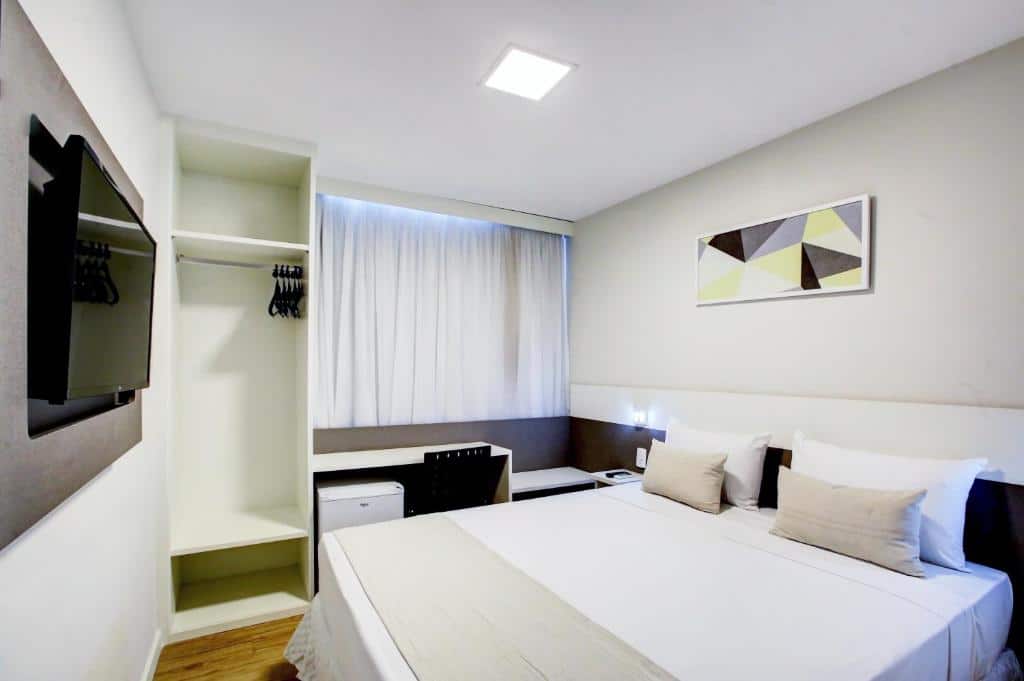 Um quarto do Go Inn Contagem - MG com uma cama de casal, TV na parede, armário aberto, escrivaninha e obras de arte abstratas acima da cama.