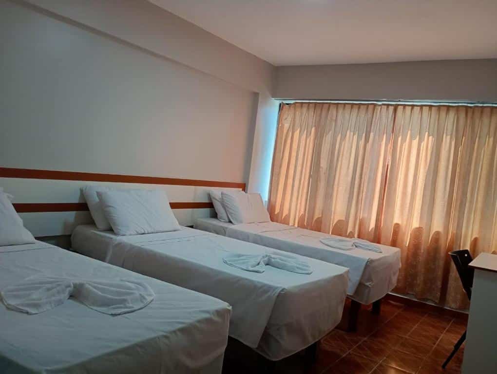 Três camas de solteiro com lençóis brancos estão dispostas em uma fileira em um quarto com uma janela coberta por cortinas bege, do Gran hotel teatro.