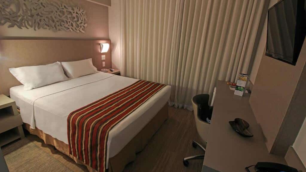 Quarto do Hotel Beaga Convention com uma cama de casal, colcha listrada, escrivaninha com cadeira, TV na parede e cortinas fechadas.