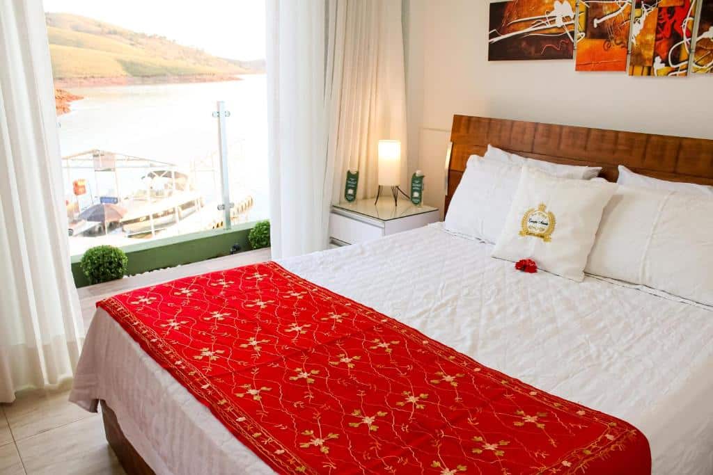 Uma cama bem arrumada com uma capa decorativa vermelha em um quarto bem iluminado com uma janela com vista para uma doca e água, do Hotel Boutique Coração Aberto.