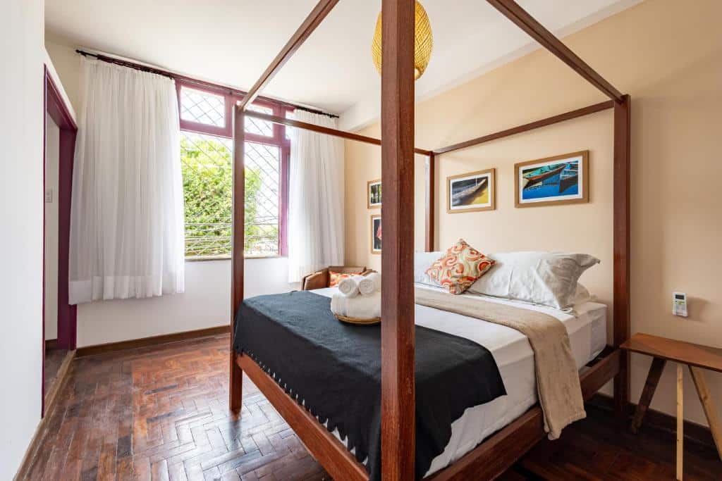 Um quarto do Hotel Casa do Bispo com uma cama de dossel, piso de madeira, quadros emoldurados na parede e uma janela com cortinas brancas.