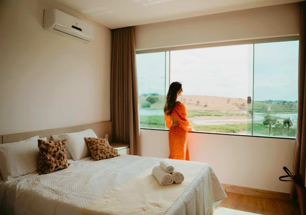 Uma mulher com um vestido laranja está ao lado de uma grande janela em um quarto, olhando para uma paisagem com água e colinas, do Hotel Chalé Capitólio.
