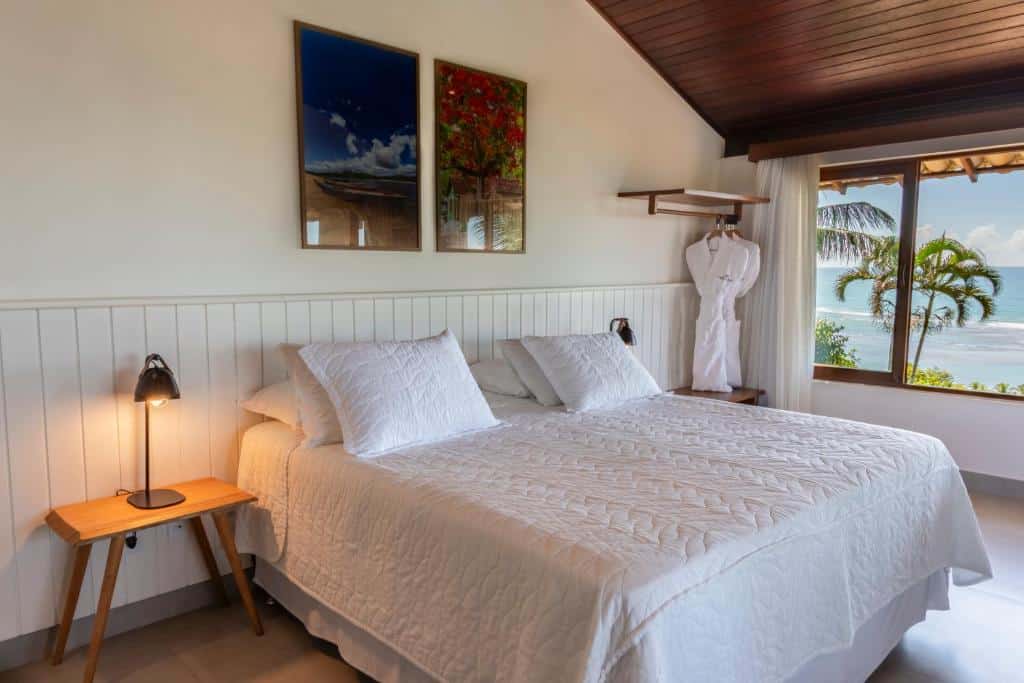 Uma cama bem arrumada do Hotel Paraiso do Morro com lençóis brancos, dois quadros acima dela e uma janela com vista para o oceano e palmeiras.