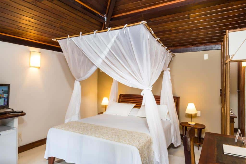 Um quarto do Hotel Pousada Coqueiros com cama de dossel, roupa de cama branca, móveis de madeira e uma televisão no lado esquerdo.