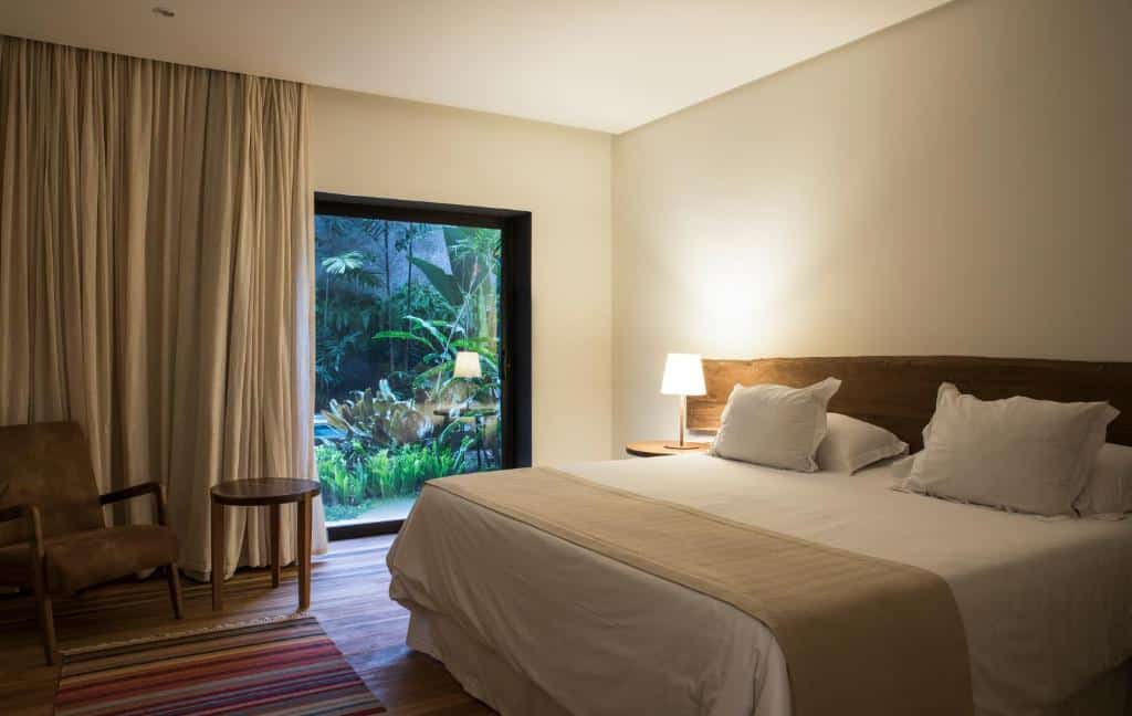 Uma cama bem arrumada em um quarto moderno com uma janela que mostra um jardim, uma poltrona, uma mesa lateral e um abajur, do Hotel Villa Amazônia.