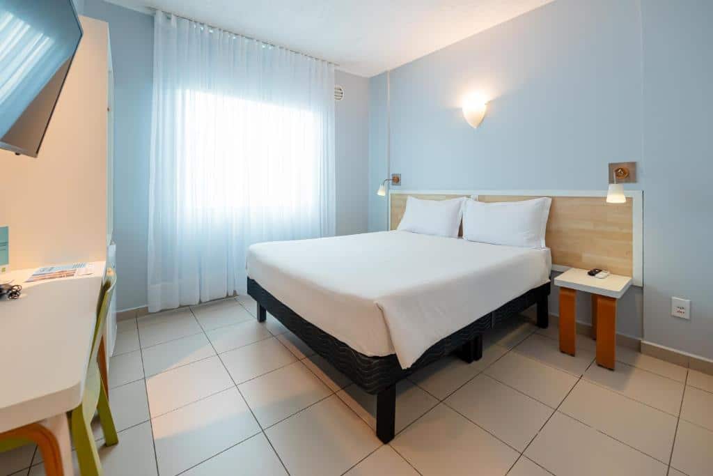Quarto do ibis Styles Manaus com uma cama de casal, duas mesinhas de cabeceira, uma luminária de parede, uma escrivaninha com uma cadeira e uma janela com cortinas transparentes.