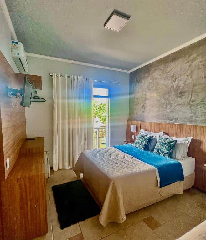 Uma cama bem arrumada em um quarto moderno com uma TV na parede, janela com cortinas e luz do sol entrando, do Lara's Hotel.