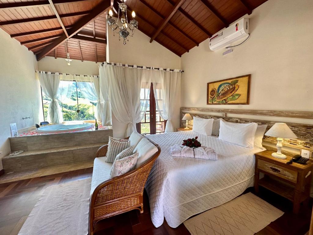 Um quarto aconchegante da Pousada Aldeia Manacás com uma cama grande, banco de vime, mesas de cabeceira e uma banheira ao lado de janelas com cortinas brancas.