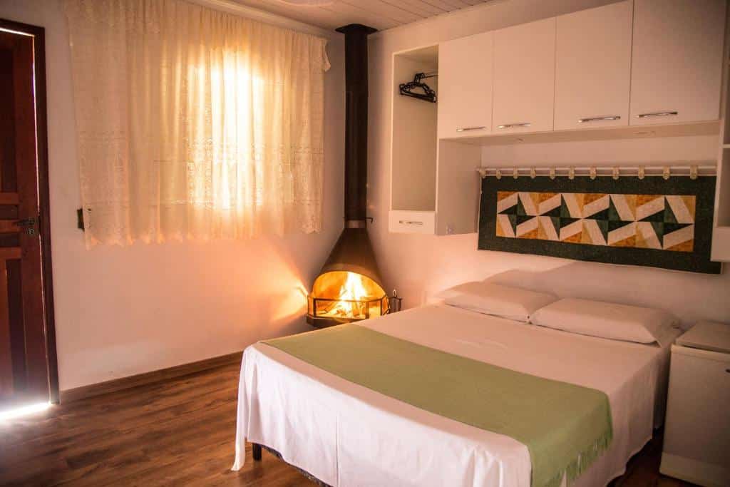 Um quarto aconchegante com uma cama de casal, uma manta verde, arte na parede, lareira, janela com cortinas e piso de madeira, da Pousada Canto Verde.