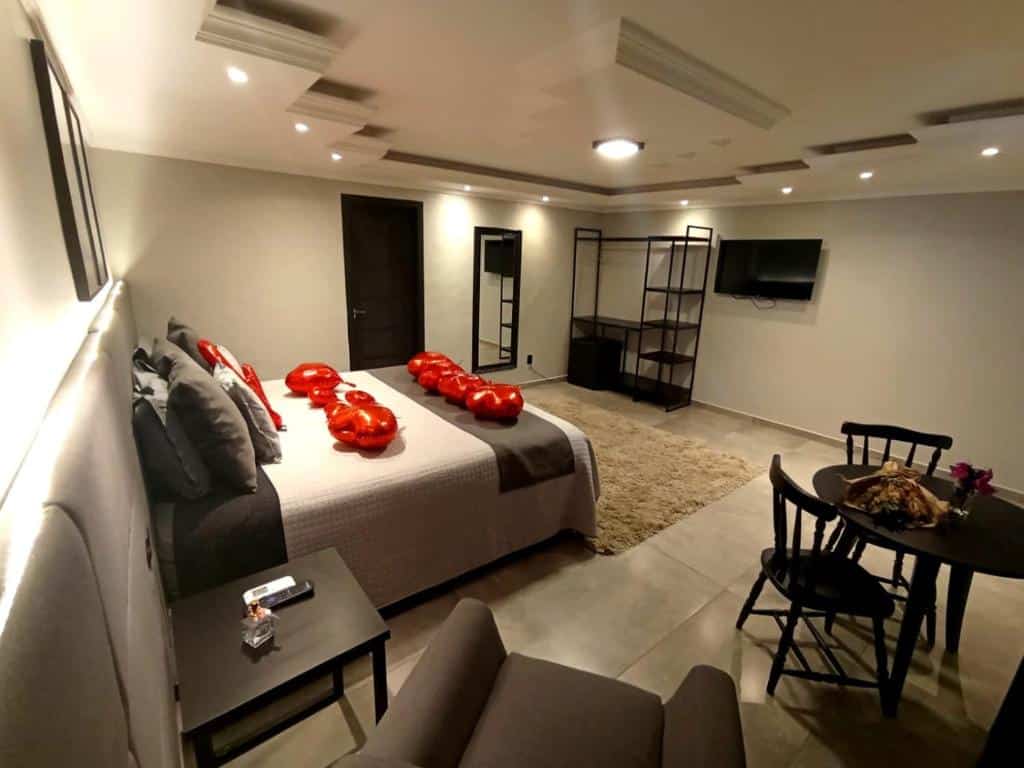 Um quarto moderno da Pousada Serra & Jardim, com uma cama de casal, balões vermelhos, uma TV, estante, poltrona, mesa redonda e decoração neutra.