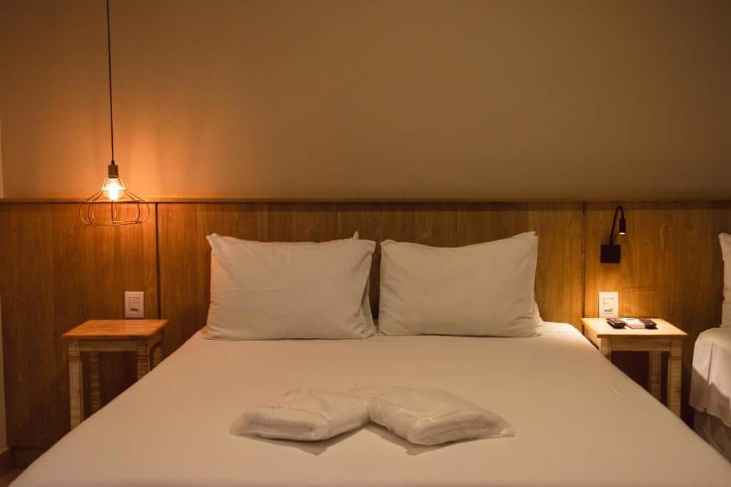 Uma cama bem arrumada com dois travesseiros brancos, toalhas dobradas e mesas de cabeceira com lâmpadas em um quarto suavemente iluminado, na Pousada Viver Capitólio.