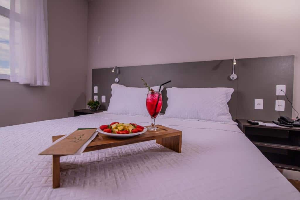 Uma cama com roupa de cama branca tem uma bandeja com um prato de frutas e um drinque vermelho com canudo e guarnição.