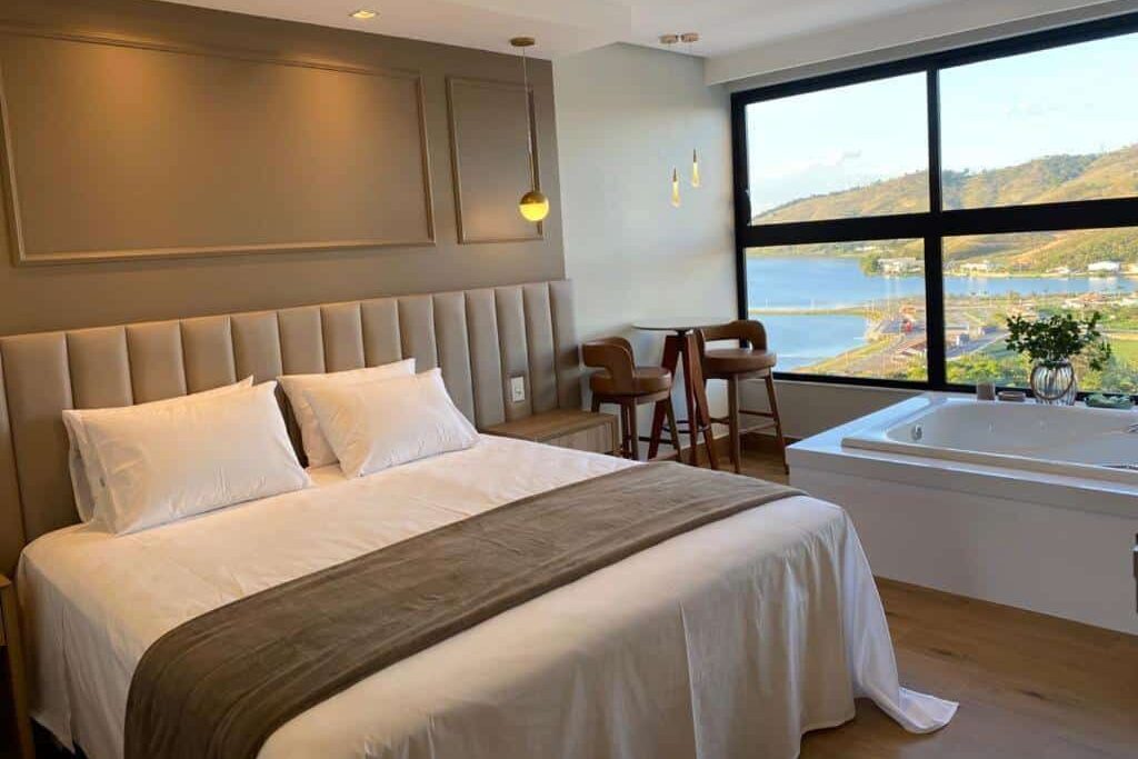 Quarto do Vista do Lago Hotel com uma cama de casal, mesa e cadeiras, banheira e janela grande com vista para a água e para a encosta.