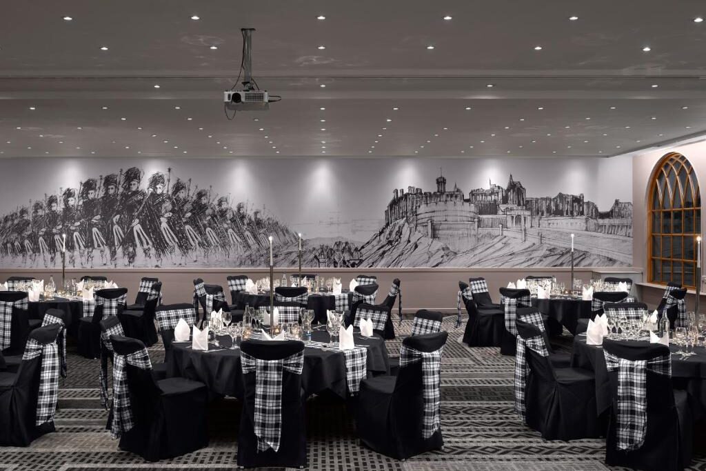 Salão de banquetes no Radisson Blu Hotel com mesas redondas, cadeiras pretas com faixas xadrez, preparado para um evento, mural de uma cena histórica na parede. Ilustra o post de onde ficar em Edimburgo.