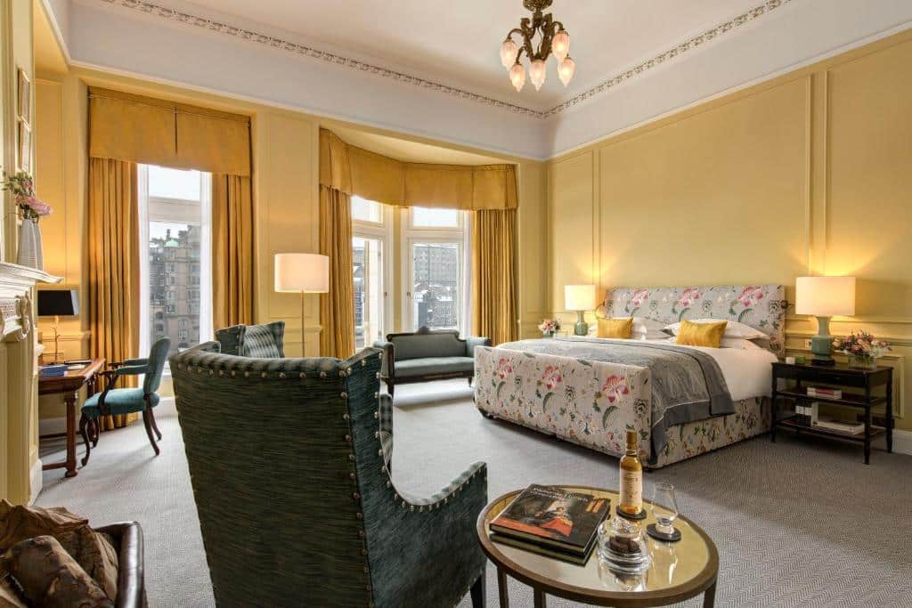 Quarto de The Balmoral Hotel espaçoso com uma cama grande, poltronas, uma escrivaninha e janelas altas com vista para a cidade e cortinas douradas. Ilustra o post de onde ficar em Edimburgo.