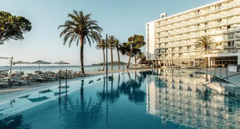 Piscina externa no The Ibiza Twiins, em Playa d'en Bossa, com espreguiçadeiras, palmeiras e um edifício de vários andares do hotel ao lado de uma praia sob um céu claro. A foto é capa do post de onde ficar em Ibiza.
