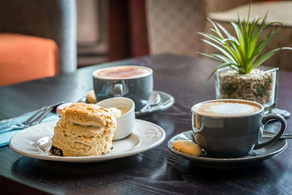 Duas xícaras de café e um scone com creme em uma mesa ao lado de um pequeno vaso de plantas no The Scholar.