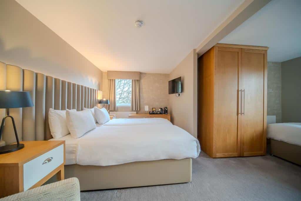 Quarto de hotel no The Scholar com duas camas grandes, um guarda-roupa de madeira, mesas de cabeceira, uma TV na parede e uma janela com cortinas claras.