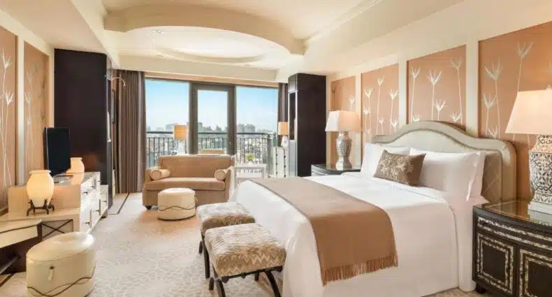 Um quarto de hotel com uma cama grande, duas banquetas, um sofá, luminárias, uma TV e uma varanda com vista para a cidade.