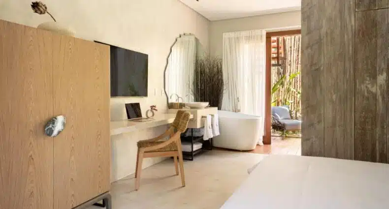 Quarto minimalista da Vila Almesca com guarda-roupa de madeira, TV na parede, escrivaninha, cadeira, espelho grande e banheira independente perto da janela. Imagem para ilustrar post sobre onde ficar em Caraíva.