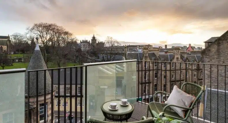 Uma pequena varanda com duas cadeiras e uma mesa tem vista para os edifícios históricos e para um parque ao pôr do sol.