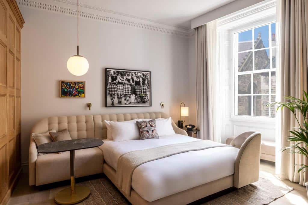 Quarto de hotel Virgin Hotels Edinburgh com uma cama grande, sofá bege, mesa redonda, arte na parede, janela grande e luz natural preenchendo o espaço. Ilustra o post de onde ficar em Edimburgo.