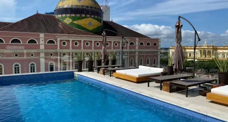 A piscina na cobertura do Juma Ópera Boutique Hotel & Spa com espreguiçadeiras tem vista para um edifício com cúpula rosa sob um céu azul com nuvens dispersas. Imagem para ilustrar post sobre onde ficar em Manaus.