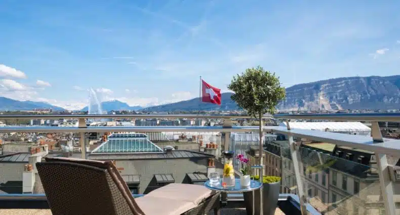 Um terraço na cobertura do Warwick Geneva com espreguiçadeira, mesa e vaso de plantas tem vista para uma paisagem urbana, montanhas e uma bandeira da Suíça. Imagem para ilustrar post onde ficar em Genebra.