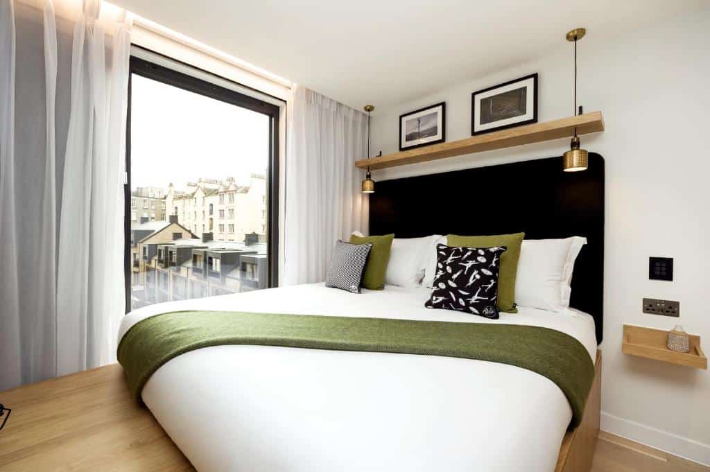 Quarto moderno no Wilde Aparthotels Edinburgh com uma cama grande, detalhes em verde, dois travesseiros, janela grande e arte emoldurada na parede acima da cabeceira. Ilustra o post de onde ficar em Edimburgo.