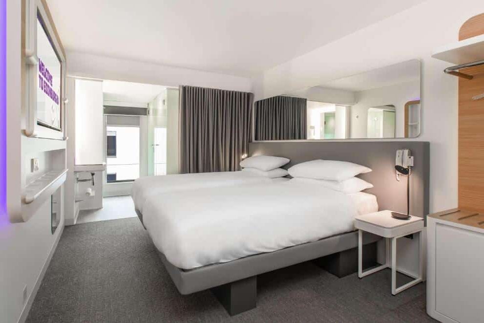 Quarto de hotel Yotel Edinburgh moderno com duas camas, TV na parede, escrivaninha, espelho grande e carpete cinza. Decoração clara e minimalista. Ilustra o post de onde ficar em Edimburgo.