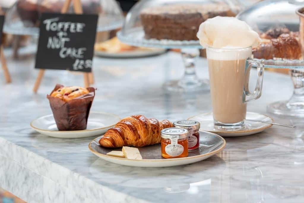 Yotel Edinburgh e Um croissant, manteiga e geleia em um prato com um muffin e um café com leite em uma mesa de mármore; bolos são visíveis ao fundo. Ilustra o post de onde ficar em Edimburgo.