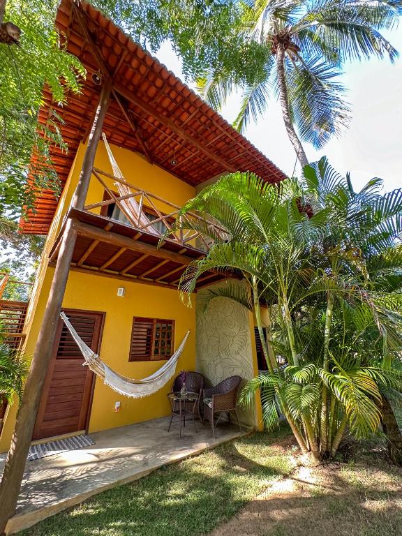 Casa amarela de dois andares da Aldeia Maracajaú com varanda de madeira, duas redes, cadeiras ao ar livre e cercada por plantas tropicais e palmeiras.