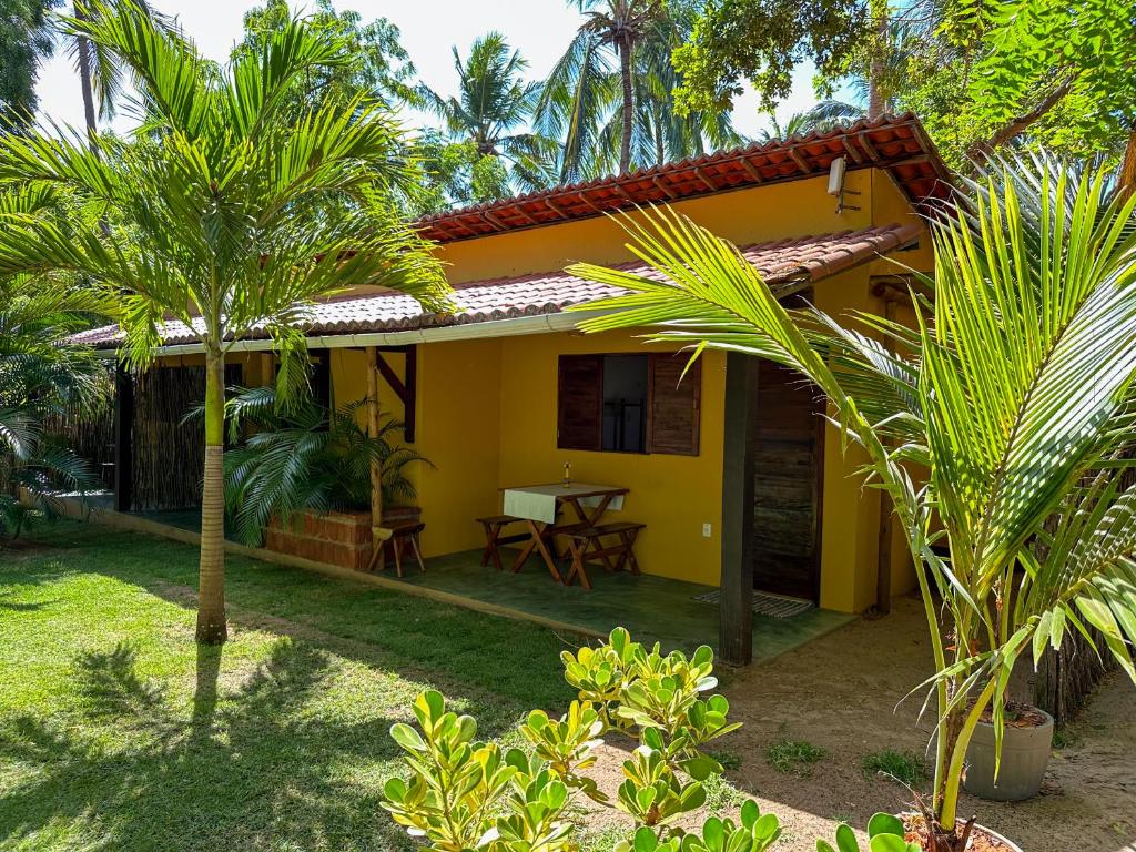 Uma pequena casa amarela com telhado de telha, mesa e cadeiras ao ar livre, cercada por palmeiras e plantas verdes. Foto da Aldeia Maracajaú camping e chalés