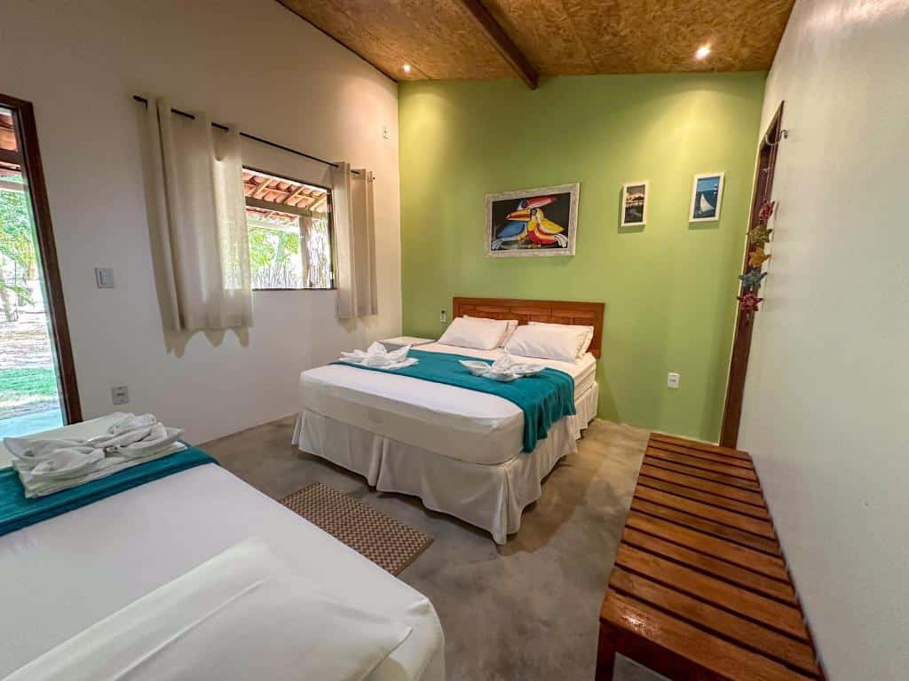 Aldeia Maracajaú Camping & Chalés. Um quarto simples com uma cama de casal e uma de solteiro, parede verde de destaque, janela, arte emoldurada e banco de madeira.