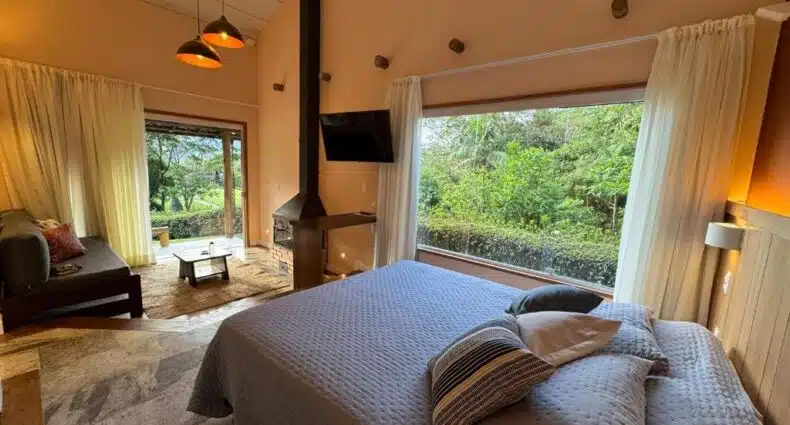 Um quarto aconchegante com uma cama grande, janela grande com vista para o jardim, TV, fogão a lenha e porta de correr para um pátio.