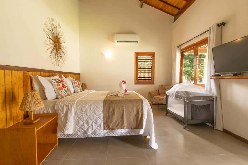Um quarto dos Bangalôs do Gameleiro com uma cama bem arrumada, uma decoração de parede, um berço, uma TV, uma poltrona e uma grande janela com cortinas. Essa é uma das indicações de onde ficar na Praia dos Carneiros.
