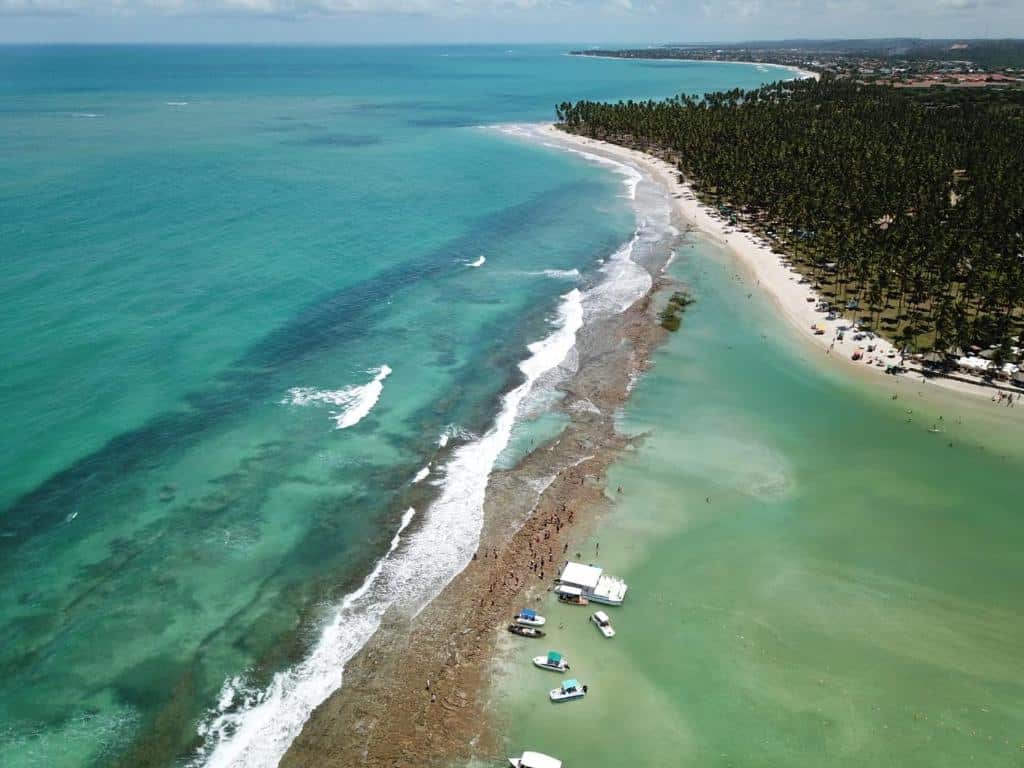 Vista aérea de uma praia tropical com água azul-turquesa clara, barcos perto da costa e palmeiras ao longo da costa.